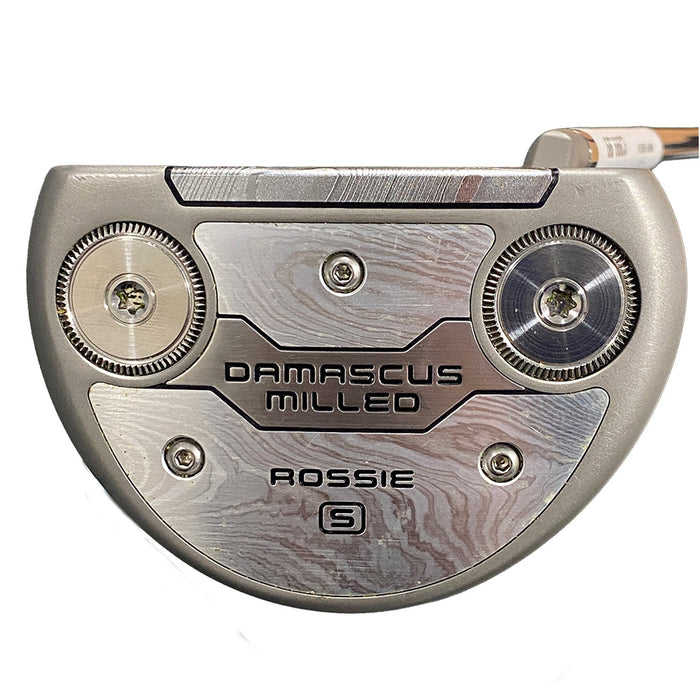 A close-up studio shot of a putter head on a golf club, featuring a brushed silver and gray metallic finish with the text 'DAMASCUS MILLED' and 'ROSSIE S' on the sole.